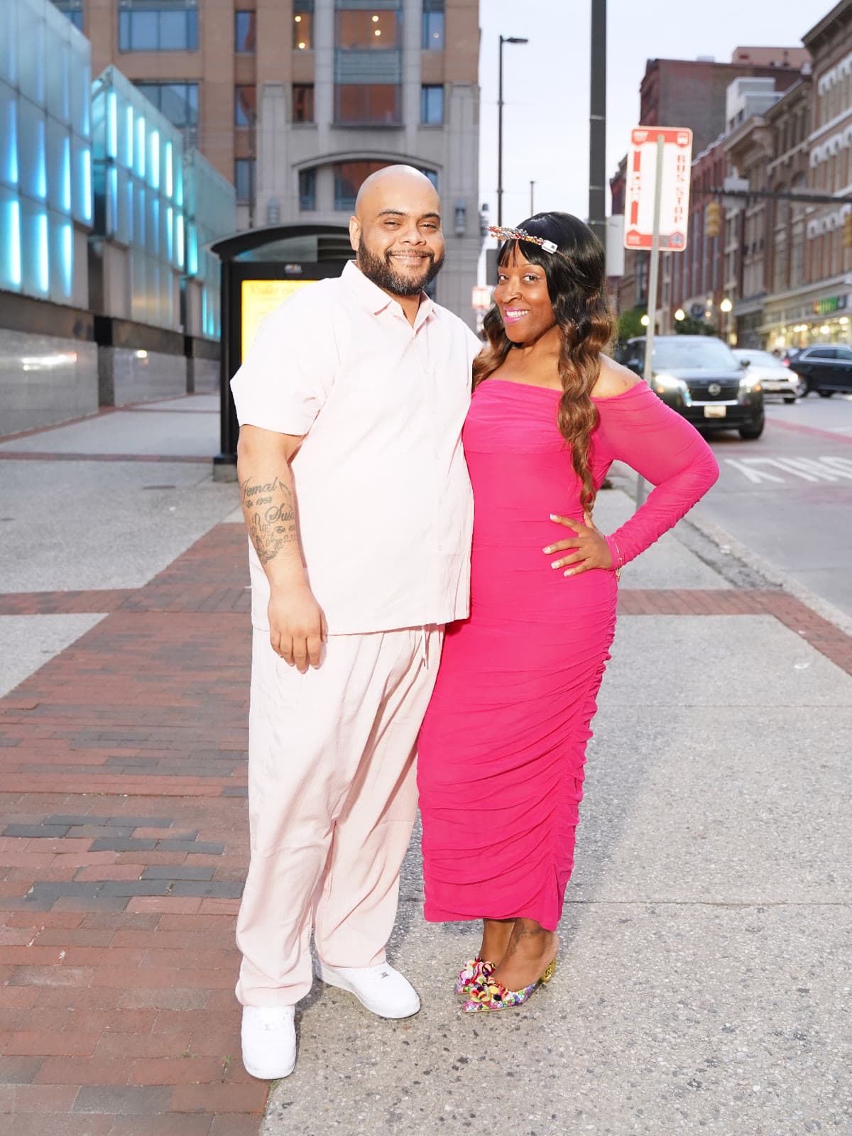 A couple dressed up for a night out, standing arm in arm on a city sidewalk at dusk
