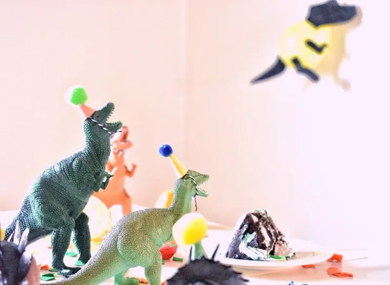 Toy dinosaurs in tiny party hats gathered around a slice of birthday cake with confetti