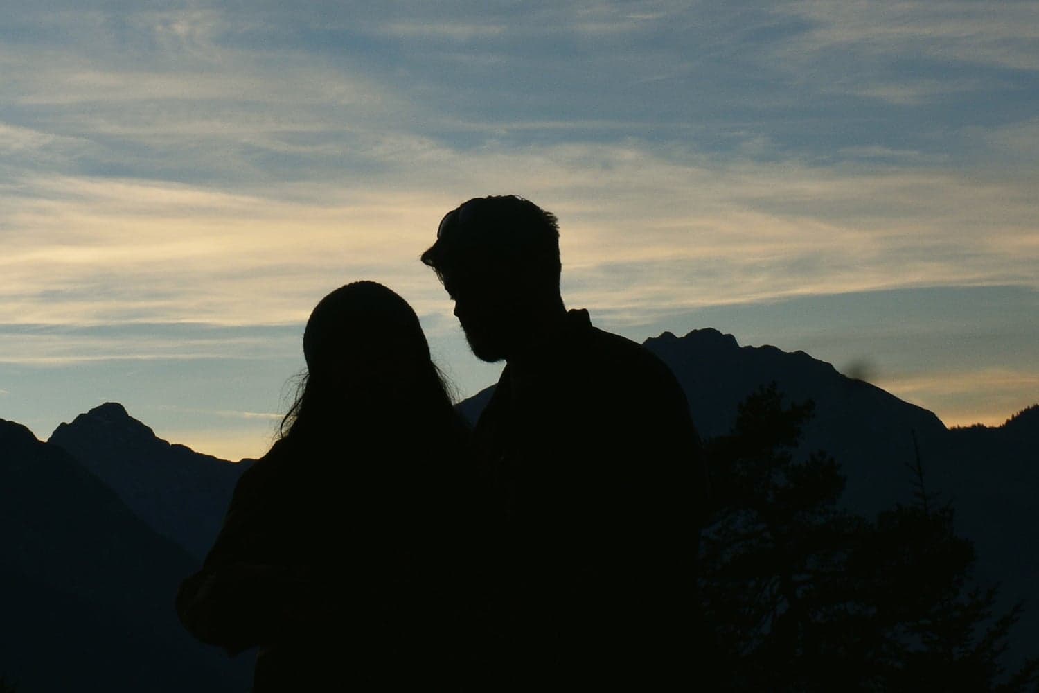 Two people standing close together in silhouette, faces almost touching, against a pale evening sky