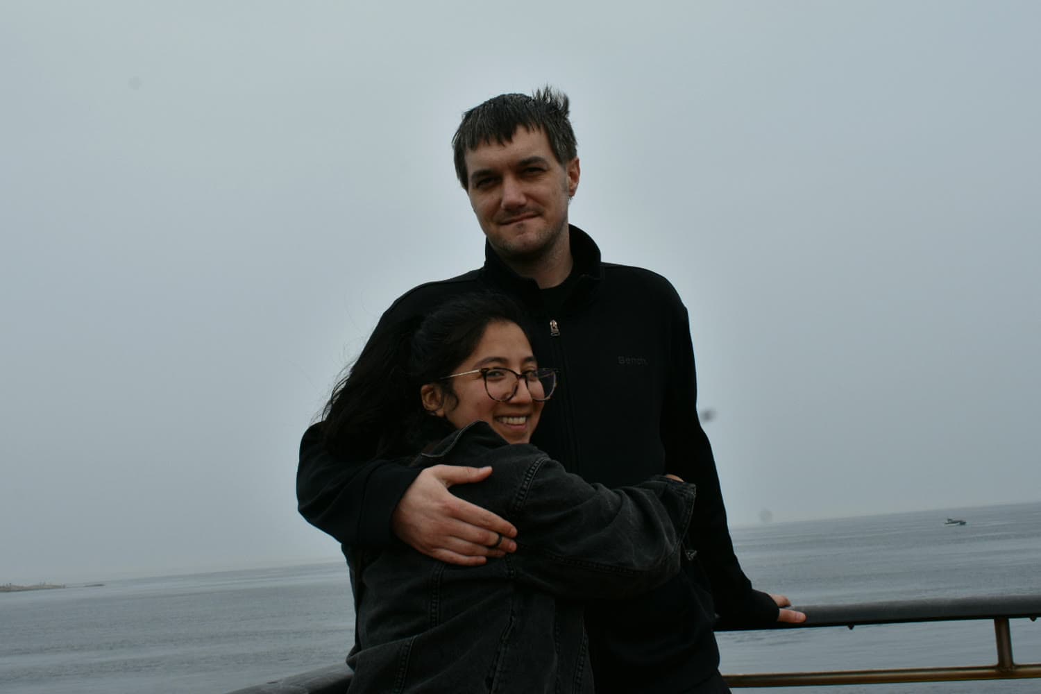 Emi and Ian on a boat deck, arms around each other, grey water and sky behind them