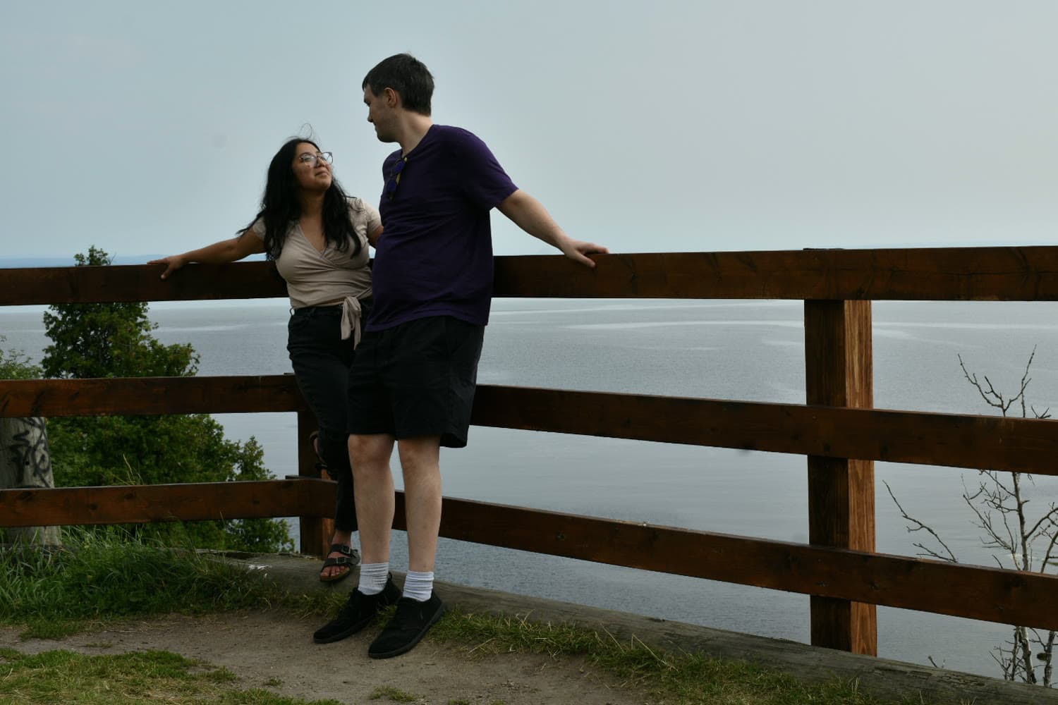 Emi and Ian standing at a wooden overlook railing above the St. Lawrence, looking at each other