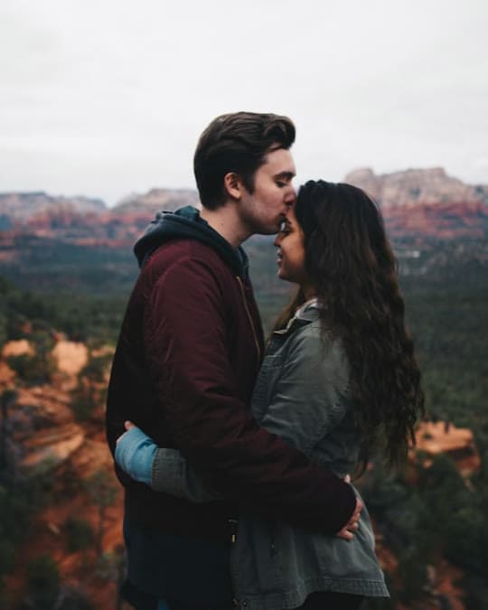 A couple embracing on a mountaintop overlook, sharing a forehead kiss