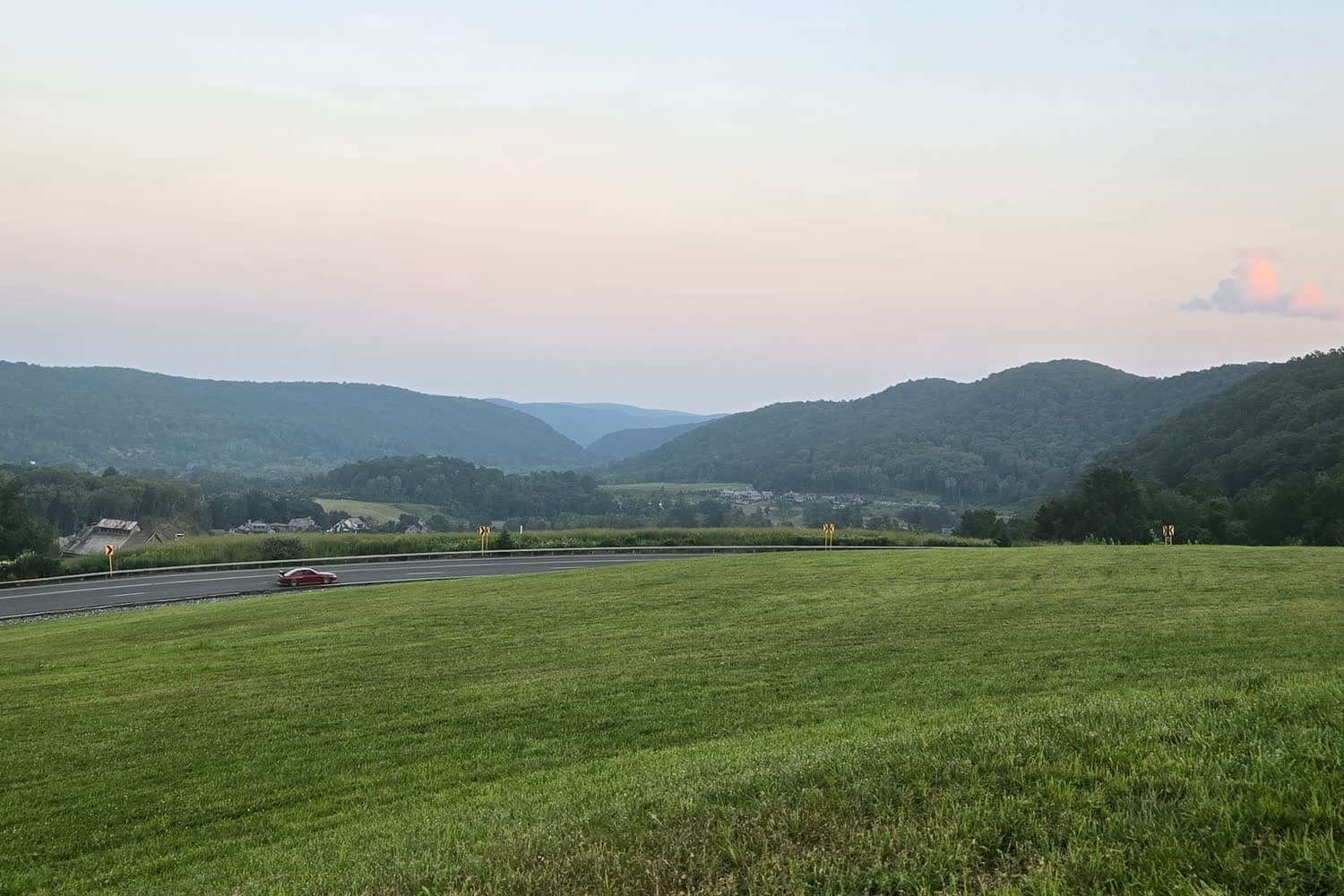 A mown green field above a curving two-lane road with one red car, wooded hills behind, under a pale pink and blue dusk sky