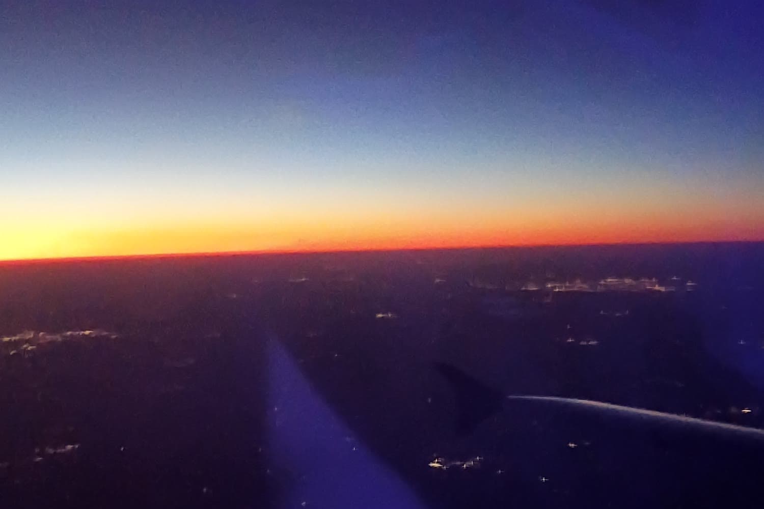 View from a plane window at dusk, an orange band on the horizon under deep blue sky, city lights below and a dark wing tip