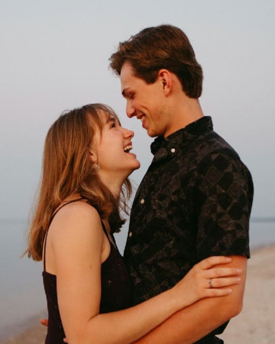 A newly engaged couple laughing together on a beach at dusk