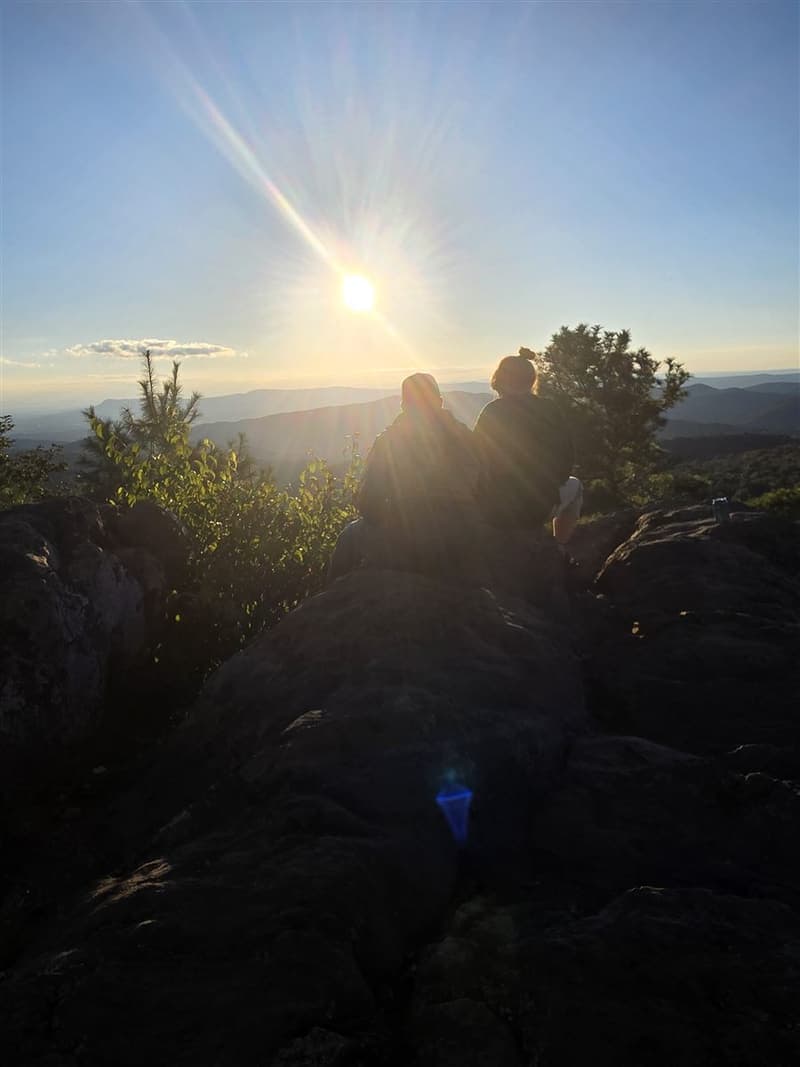 A couple sitting on a rocky summit watching the sun set over the mountains