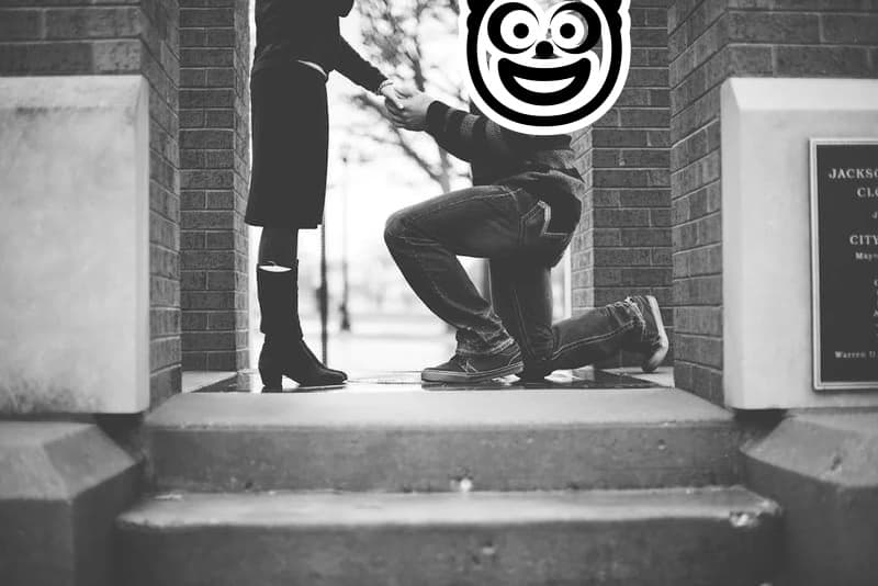 A black and white photo of a man proposing on one knee, with a cartoon clown face drawn where his head would be