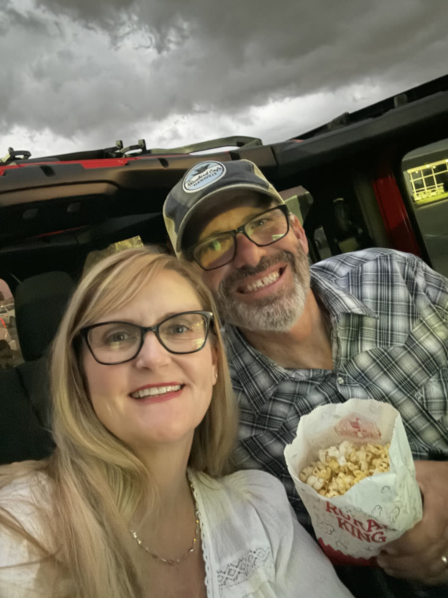 Shay and Brett smiling in their Jeep with a bag of popcorn under a cloudy evening sky